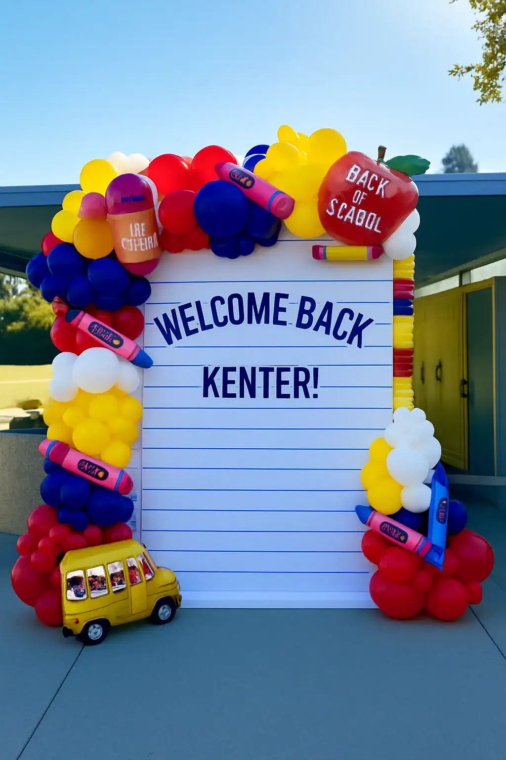 A “Welcome Back Kenter!” sign decorated by a balloon artist with colorful balloons, giant crayons, an apple, and a yellow toy school bus, set outdoors by a building.