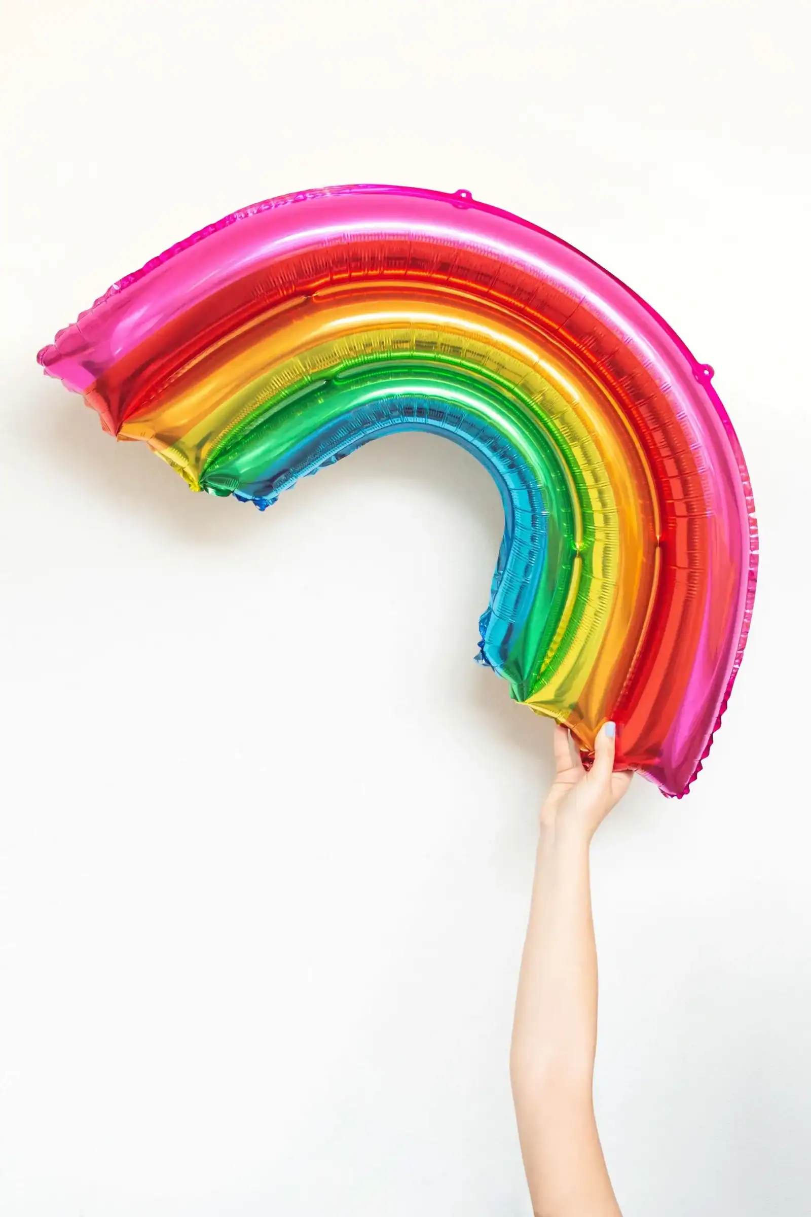 A hand holds up a metallic rainbow-shaped balloon against a plain white background, showcasing the playful work of a talented balloon artist from Los Angeles.