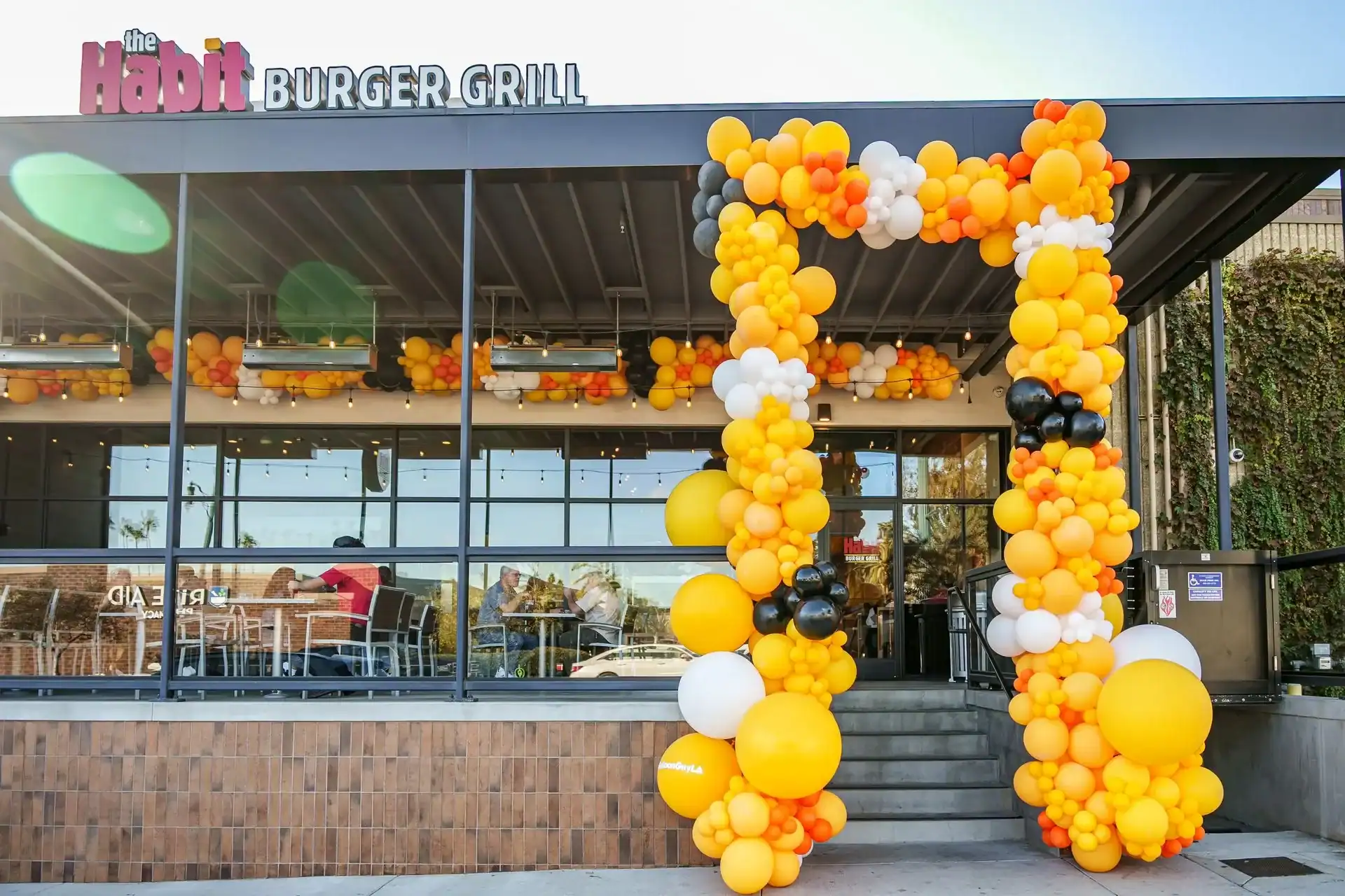 The Habit Burger Grill storefront features vibrant balloon decor, with an arch of yellow, orange, white, and black balloons welcoming guests near the entrance.
