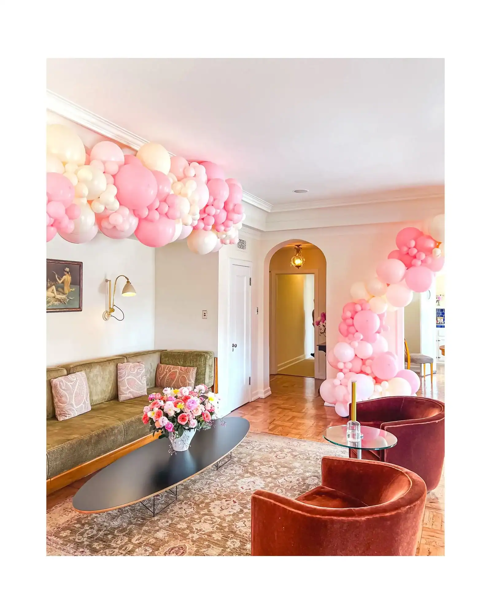 A living room decorated with balloon installations in clusters of pink and white, featuring a green couch, two rust-colored chairs, a coffee table with flowers, and a patterned rug.
