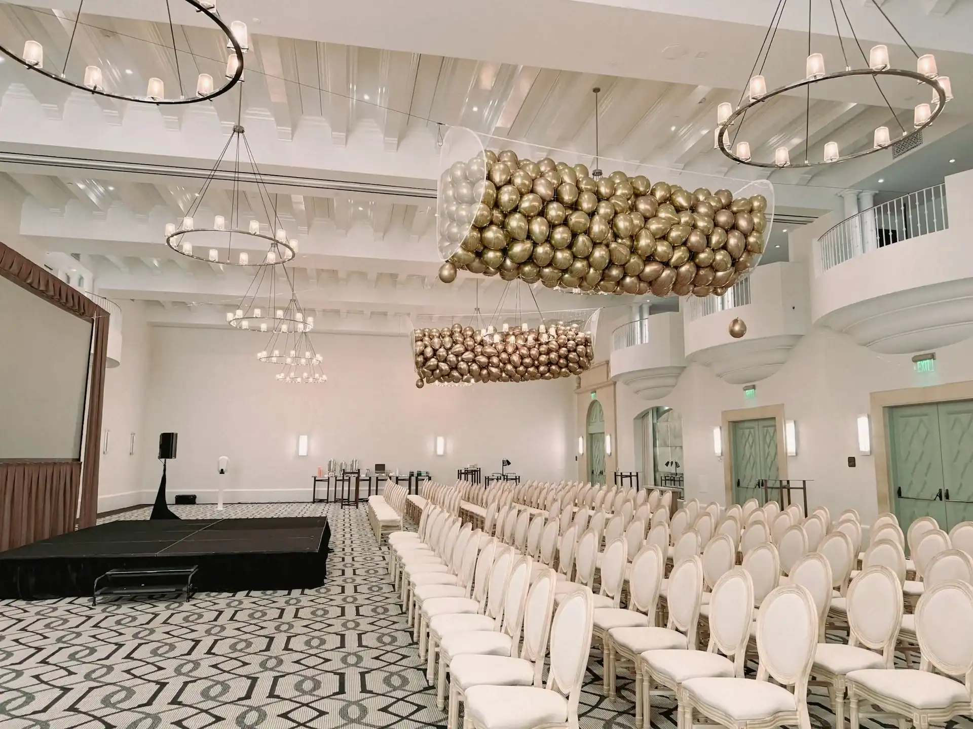 A spacious, elegant event hall in Los Angeles with rows of white chairs, a raised stage, stunning balloon decor, and modern chandeliers hanging from the ceiling—perfect for your next corporate event.