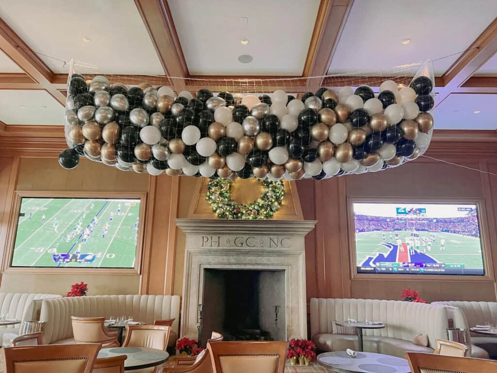A large net filled with black, white, gold, and silver balloons hangs from the ceiling above a lounge area—perfect for a corporate event in Los Angeles—with two TVs showing football games and a wreath above the fireplace.