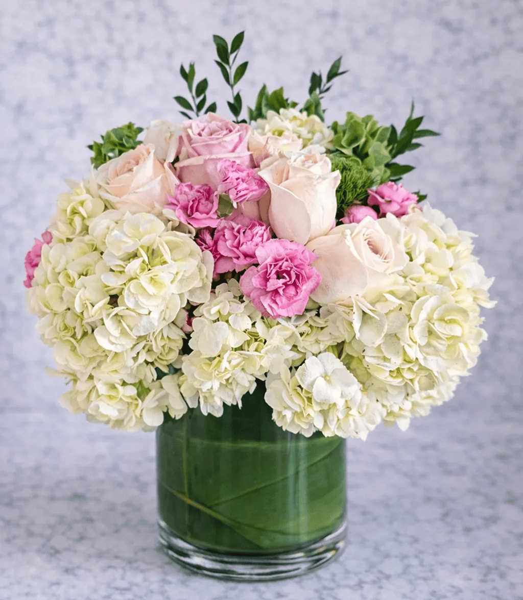 A glass vase filled with white hydrangeas, light pink roses, pink carnations, and green leaves is arranged against a light, marbled background—perfect for complementing elegant balloon decor in Los Angeles.