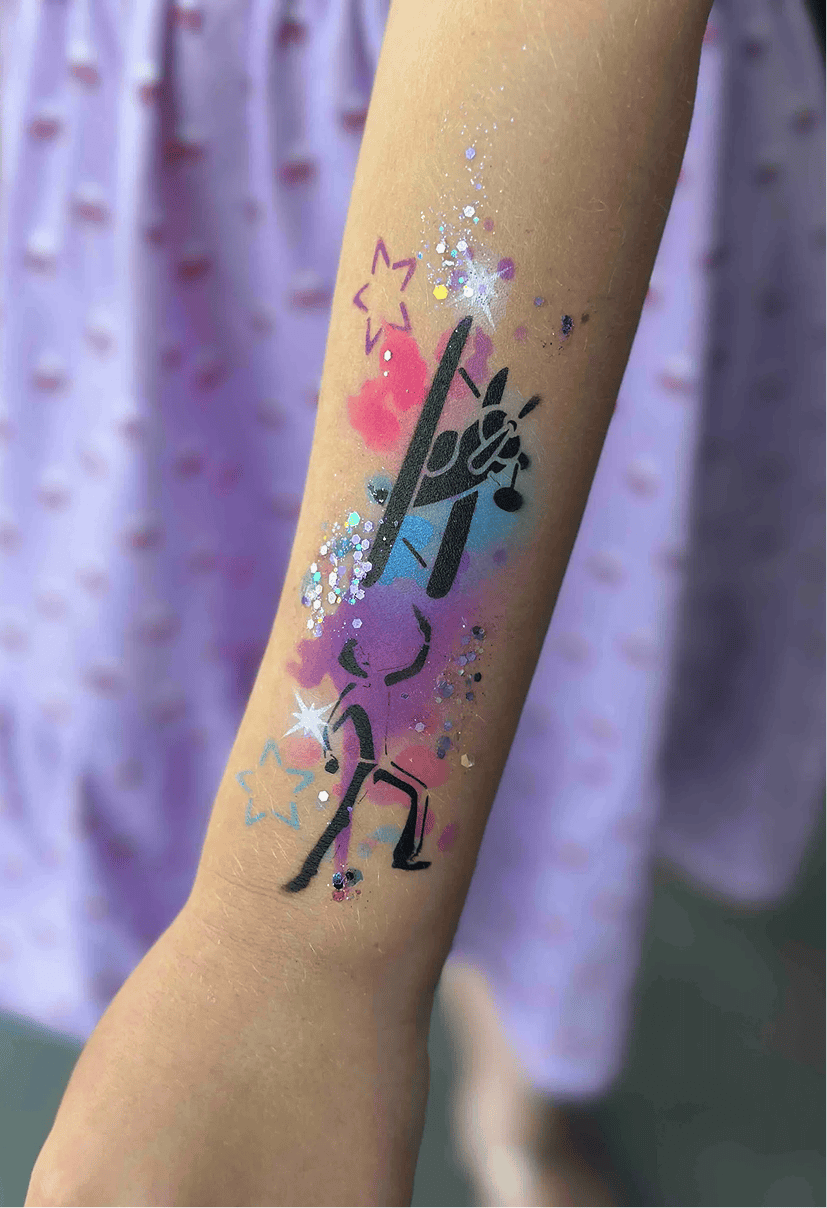 A child’s forearm with a painted design of a black dancer on a colorful, glittery background with stars, balloons nearby, against a blurred purple skirt at a lively Los Angeles event.