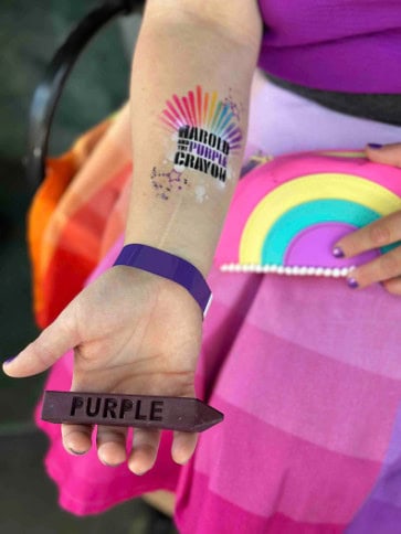 A person in Los Angeles holds a large purple crayon labeled "PURPLE." Their arm has a colorful tattoo reading "HAROLD AND THE PURPLE CRAYON." They wear a purple wristband and hold a rainbow purse, surrounded by vibrant balloon decor.