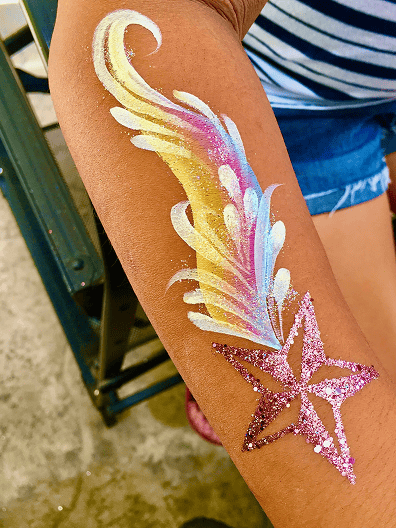 A child's forearm features a painted design of a glittery pink star with a yellow, white, and blue comet tail, captured against the backdrop of Los Angeles as a balloon artist creates vibrant balloons nearby.