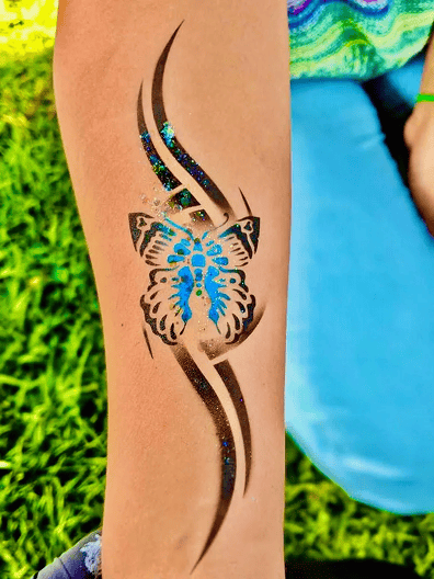 A forearm features a temporary tattoo of a blue and black butterfly with abstract black and blue dotted lines, set against grass, blue pants, and vibrant balloon decor reminiscent of Los Angeles celebrations.