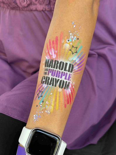 A forearm with a "Harold and the Purple Crayon" temporary tattoo, colorful rays, star outlines, and glitter accents; person wearing a white watch and purple clothing at a corporate event with creative balloon decor.