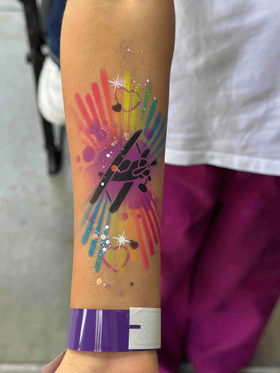 A forearm displays a colorful painted design with rainbow handprints, black geometric shapes, glitter, and a purple wristband, perfect for pairing with vibrant balloon decor at any festive event.