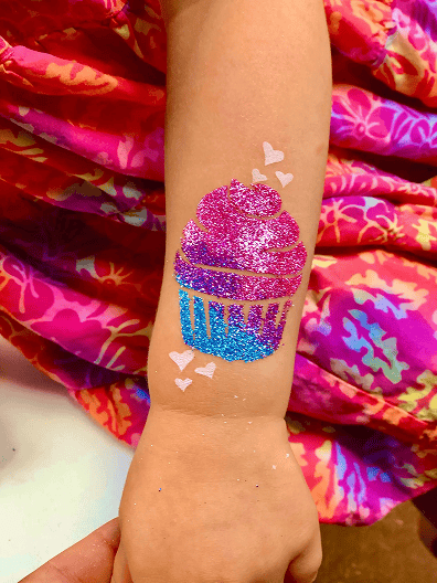 A child's arm with a glittery cupcake tattoo in pink and blue, surrounded by small white heart designs, against a colorful patterned skirt at a Los Angeles balloon artist’s party.