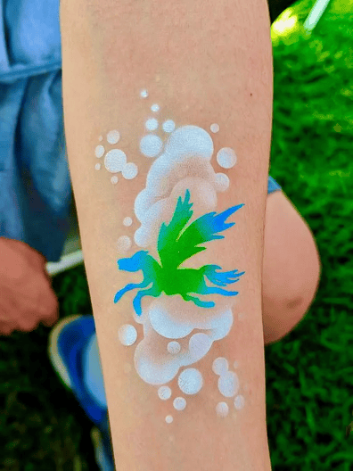 A green and blue fairy with wings is painted on a forearm surrounded by white bubble designs; perfect inspiration for corporate event face painting ideas in Los Angeles. Grass and a person's knee are visible in the background.