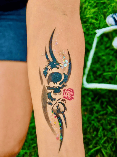 A forearm tattoo featuring a black tribal skull design with a red rose and glitter accents, shown outdoors on grass—perfect inspiration for creative balloon decor or bold corporate event themes.