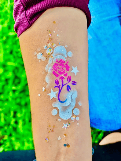 A forearm displays a painted design featuring a pink rose with a blue stem, surrounded by white stars, circles, glittery accents, and whimsical balloons for added festive flair.
