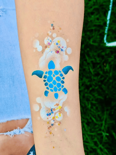A blue sea turtle design with white bubbles and colorful glitter is painted on a person’s forearm, perfect for pairing with vibrant balloon installations by a skilled balloon artist.