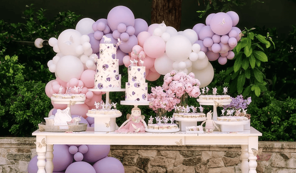 A dessert table decorated with stunning balloon installations in purple and pink, tiered cakes, flowers, and assorted sweets set outdoors against greenery and a stone wall.