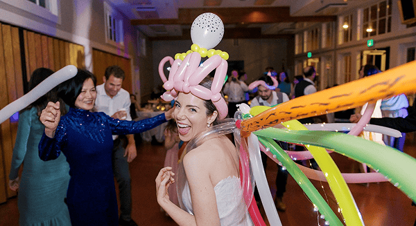 A woman in a white dress smiles while wearing a balloon hat, surrounded by vibrant balloon decor and colorful decorations at an indoor party with other people dancing around her.