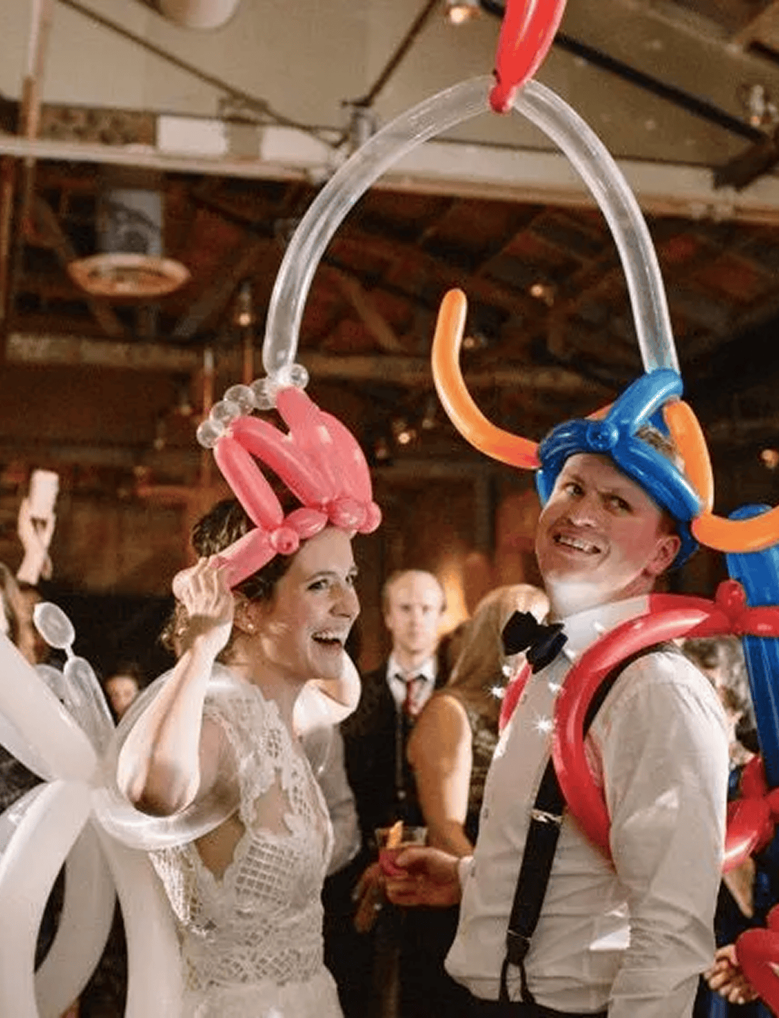 A bride and groom at a Los Angeles wedding reception wear colorful balloon hats, created by a talented balloon artist, while smiling and standing among guests.