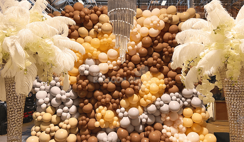 A decorative balloon decor backdrop made of clustered brown, tan, cream, and white balloons stands between two ornate feather arrangements with crystal bases—perfect for elevating any corporate event.