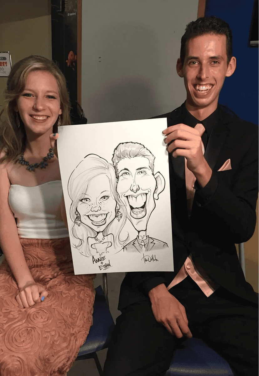 A smiling woman and man sit side by side in formal attire while the man holds a caricature drawing of them, surrounded by vibrant balloon decor.
