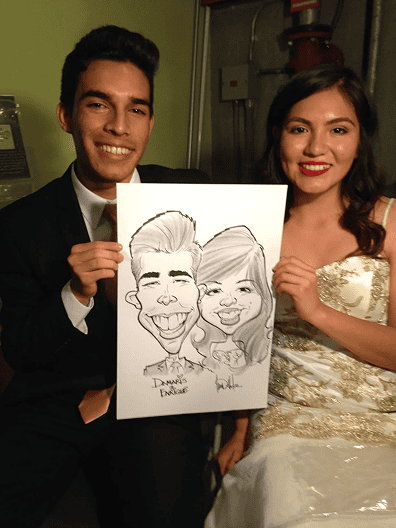 A young man and woman in formal attire sit together, smiling and holding a caricature drawing of themselves at a lively corporate event in Los Angeles.