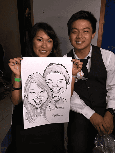 A woman and man sit together smiling, while the woman holds a caricature portrait of them both, surrounded by colorful balloon decor.