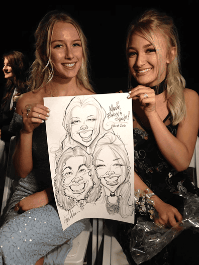 Two women sitting and smiling while holding a caricature drawing of themselves at a corporate event, surrounded by festive balloon decor.