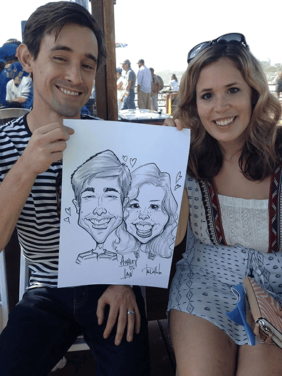 A man and woman sit side by side outdoors in Los Angeles, smiling and holding up a black-and-white caricature drawing of themselves.