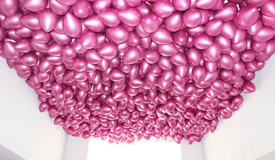 A large number of shiny pink balloons are clustered together on the ceiling of a white indoor space, creating stunning balloon decor.