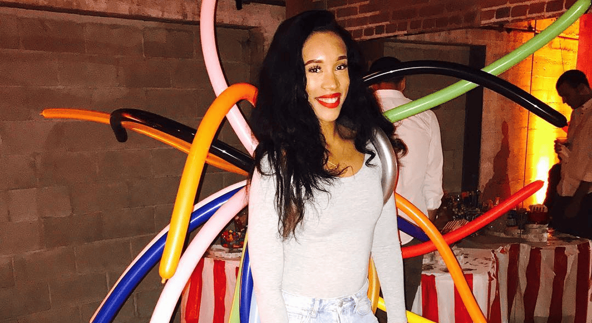 A woman in a light gray long-sleeve top and jeans stands smiling in front of vibrant balloon decor created by a talented balloon artist at a festive indoor corporate event.