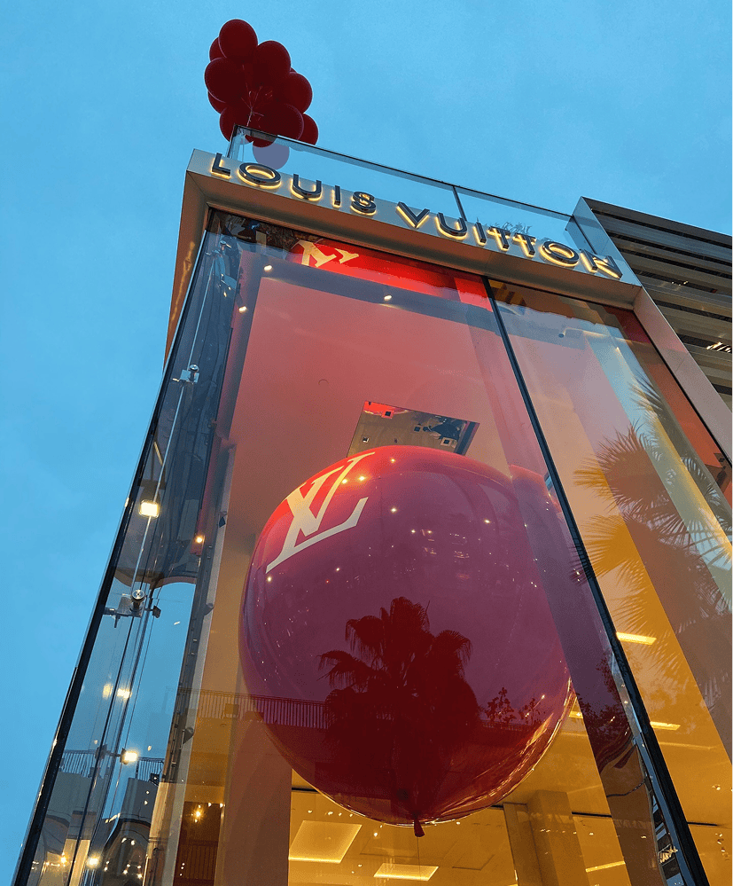 A large red LV balloon and smaller red balloons, crafted by a skilled balloon artist, are displayed at the glass entrance of a Louis Vuitton store at dusk.