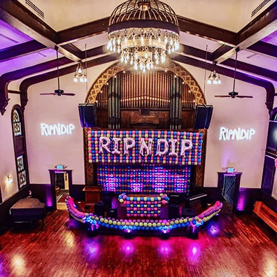 A decorated event venue with wooden floors, vibrant balloon installations on the stage, "RIPNDIP" signs on the wall, and a chandelier overhead.