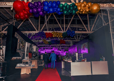 A decorated indoor event space with a red carpet, vibrant balloon installations, food stations, and people walking inside—perfect for a memorable corporate event.