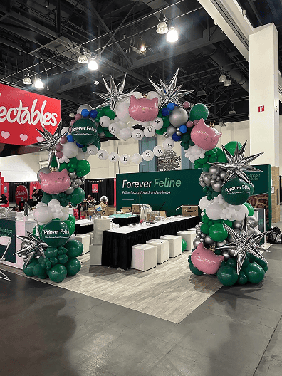 Balloon arch with metallic stars and cat designs frames the entrance to a "Forever Feline" booth at a corporate event. Balloon installations by a skilled balloon artist add flair, while white stools and tables are visible inside the booth.
