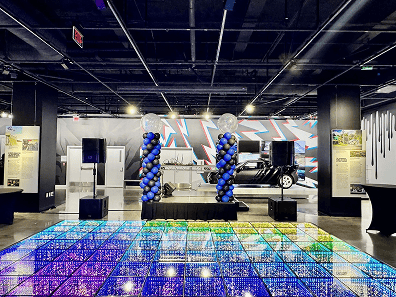 A modern event space with a lit-up glass dance floor, balloon artist installations, a stage with speakers, and a car display set against a colorful mural wall.