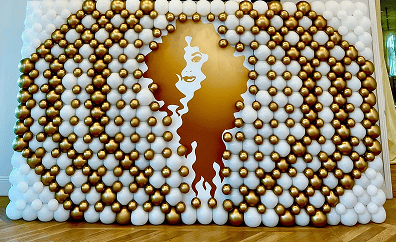 A decorative wall made of white and gold balloons features a cutout silhouette of a woman's face and hair in the center, showcasing elegant balloon decor.