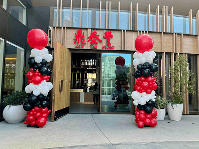 Entrance of Din Tai Fung restaurant decorated with stunning balloon decor featuring tall columns of red, white, and black balloons, accented by large potted plants on either side.