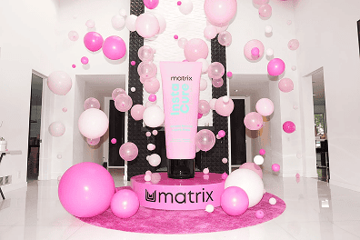 A large pink Matrix hair product tube is displayed on a circular platform surrounded by chic balloon decor in a bright, modern room—perfect inspiration for a stylish Los Angeles corporate event.