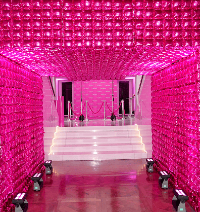 A hallway lined with shiny pink balloon decor leads to white stairs with pink-lit walls and velvet ropes at the top.
