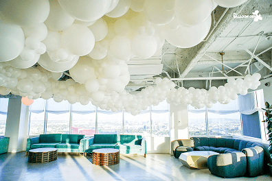 A modern lounge with blue sofas, round tables, and large windows, featuring elegant balloon decor with clusters of white balloons hanging from the ceiling.