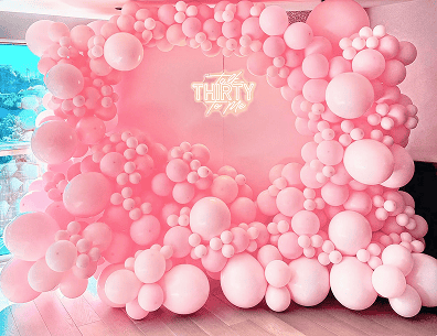 A large pink balloon arch frames a neon sign that reads "Talk Thirty To Me," set up indoors with stylish balloons for a 30th birthday celebration in Los Angeles.