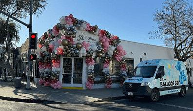 A storefront in Los Angeles is decorated with pink, white, and gold balloon decor; a van labeled "The Balloon Guy," a local balloon artist, is parked outside on a sunny street corner.