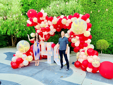 Two people stand in front of a decorative balloon arch with red, white, and gold balloons at a corporate event outdoors in Los Angeles, surrounded by trees and greenery.