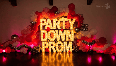Large illuminated letters spelling "PARTY DOWN PROM" are surrounded by vibrant balloon installations in red, white, gold, and silver, adding a festive touch perfect for any corporate event.