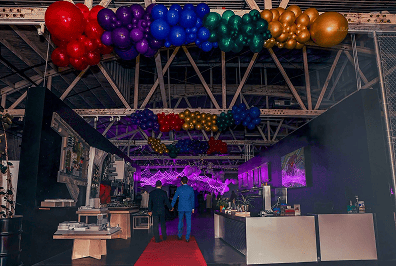 Two people walk down a red carpet into a decorated venue, where stunning balloon decor and purple lighting set the mood for this corporate event, with colorful balloons overhead.