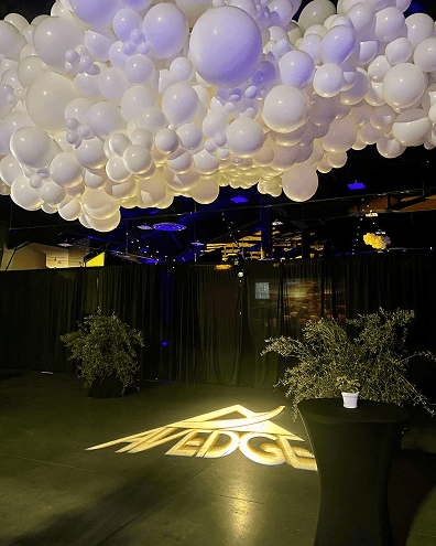 Large cluster of white balloon decor suspended from the ceiling, with the word "EDGE" illuminated on the floor below; perfect for a corporate event, with two small potted plants and a cocktail table nearby.