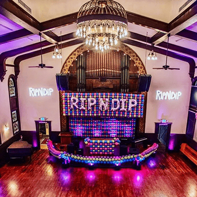 A decorated indoor venue with striking balloon installations, a large "RIPNDIP" sign made of colorful balloons above a stage, purple lighting, and a chandelier hanging from the ceiling.