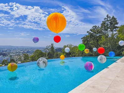 Colorful balloon installations float above an outdoor infinity pool, with city buildings and green trees visible in the background under a partly cloudy sky.