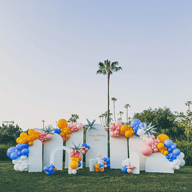 Outdoor birthday party setup with white display boards, vibrant balloon installations in blue, yellow, pink, and white, and palm trees in the background.