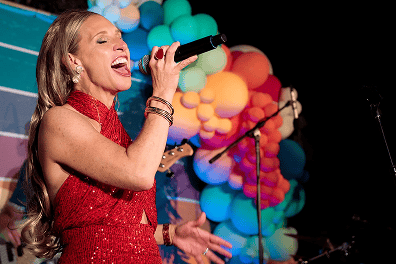 A woman in a red dress sings into a microphone on stage at a corporate event in Los Angeles, with vibrant balloon installations in the background.