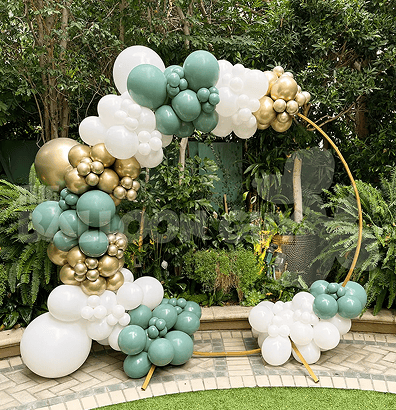 A gold circular frame is decorated with clusters of white, green, and gold balloons, creating a stunning backdrop for a corporate event set outdoors among lush green plants in Los Angeles.